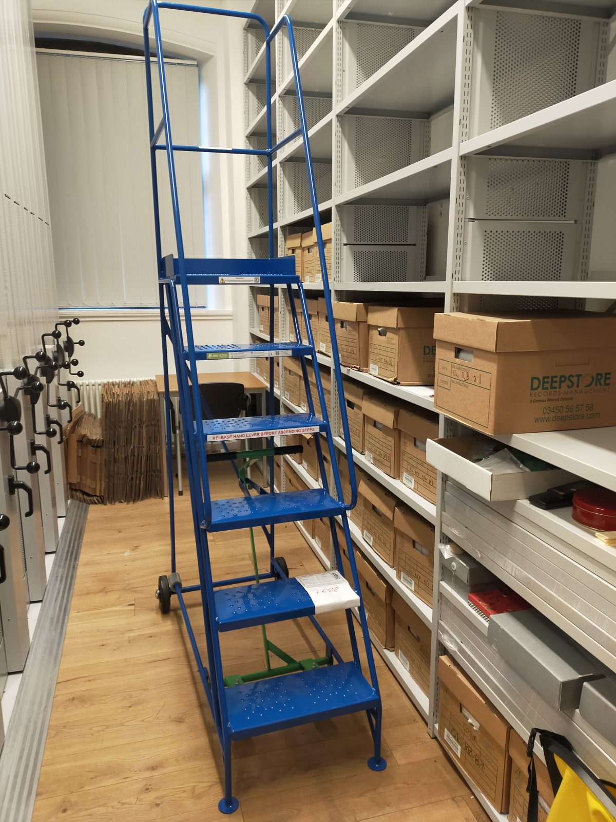 View of a library floor, with boxes on the shelves, and a tall blue ladder in the foreground.