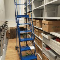 View of a library floor, with boxes on the shelves, and a tall blue ladder in the foreground.