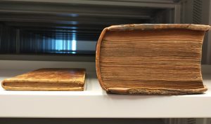 Two old books side by side on a shelf, one very small and thin (on left) and one very large and fat (on right)